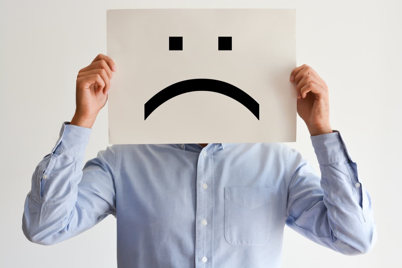 A man in an office shirt holding a sheet of paper with a frowning face drawn on it in front of his own face