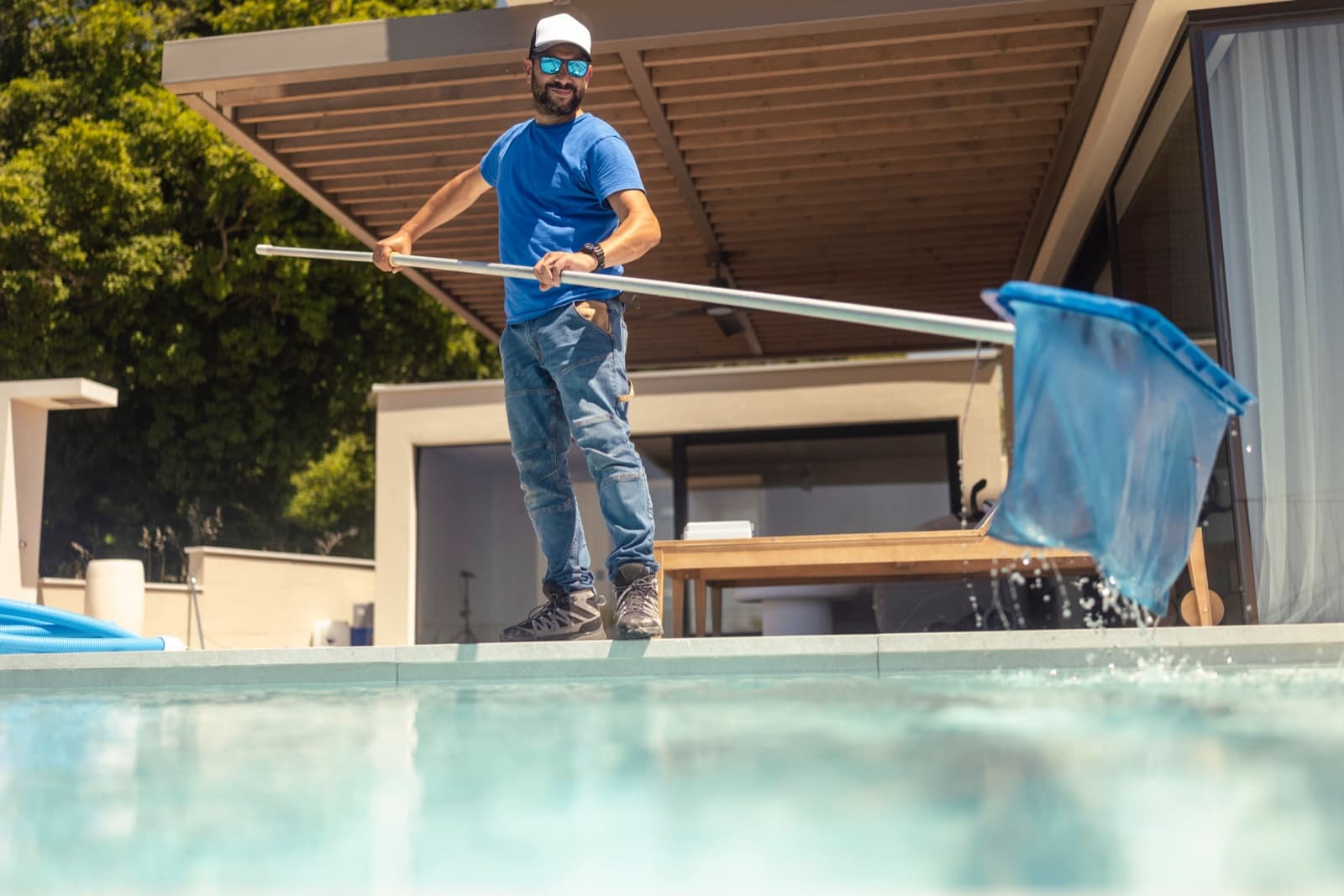 A pool professional in a cap and sunglasses lifting a skimmer net from a backyard pool, photographed from water level